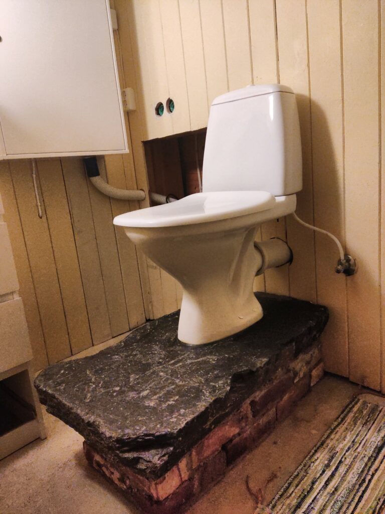 A toilet raised a 2 layers of brick and standing on a great slab of slate.